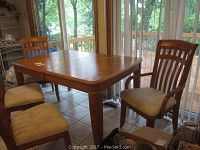 Wooden dining table with four chairs in natural wood finish, two chairs with armrests, two without, beige stained upholstery.