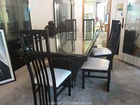 Photo showing the black lacquered dining table with glass top and four high-back chairs around it, white cushions visible on seats.