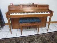 Front view of the Wurlitzer piano and bench showing the keys, legs, and cushioned bench seat with a tiled floor and a rug in the foreground.
