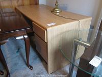 Full side view of light wood veneer office desk with 4 drawers on right side. Desk surface has a brass lamp on top.
