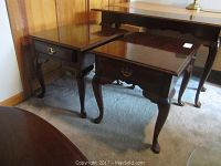 Two matching wooden end tables with carved cabriole legs, each with a drawer and brass handle, positioned near a larger table against wood-paneled wall.