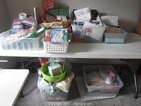 Wide shot of a table covered with multiple plastic bins and containers filled with a variety of crafting supplies including photo albums, cards, and ribbons.
