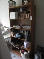 Front view of 5-tier particle board bookcase containing various household items including boxes, picture frames, and baskets.