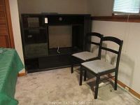 Black entertainment center with glass doors and two matching wooden chairs with cushioned seats, positioned in a carpeted room.