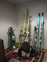 Shows five sets of skis standing against the wall with ski poles, a green golf bag with clubs, and a box with hand weights on the floor.