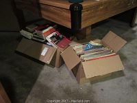 Two open cardboard boxes on the floor; one filled with assorted books and magazines, the other with vinyl records.