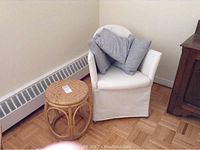 White fabric-covered Ikea chair with three grey throw pillows inside, on wooden floor next to baseboard heater and wooden furniture
