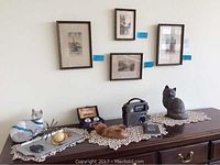 Four framed watercolor art pieces on wall above wooden dresser with cat figurines, vintage radio and other small decor