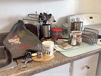Countertop with stainless steel cutlery in holder, assorted kitchen items, glass canisters, and Hello Kitty apron
