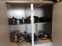 Kitchen cabinet shelf with multiple stainless steel saucepans, a double boiler, and frying pans with some lids.