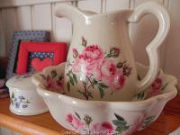 Close-up of decorative wash basin and matching rose-patterned pitcher by Cornwall Kennewick Pottery, England, with nearby small ceramic floral lidded container and decor frames on shelf.