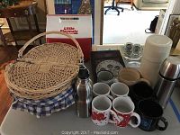 View showing medium round wicker picnic basket with blue and white checkered fabric, Igloo cooler, white and stainless steel thermoses, coffee mugs, trivets, ashtrays, and shot glasses on table