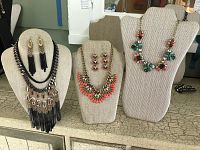 Three necklace displays showing two sets of necklace and earring pairs: one floral multicolor glass stone necklace with matching earrings, one black fringe tassel necklace with matching tassel earrings.
