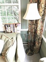 Floor lamp standing next to couch showing brass base, glass tray, and cloth shade with wooden magazine rack behind couch.