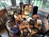 Overview of kitchen appliances and tools on round wooden table including blender, coffee grinders, sandwich toaster, popcorn popper, coffee pots, oven mitts and wooden cutting boards.