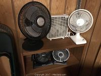 Five desk fans arranged on wooden shelves showing different brands and sizes including Lasko, Holmes, Blizzard, Duracraft and BreezeN fans.
