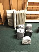Overview of six heaters and one humidifier arranged on carpet floor, showing various sizes and brands.