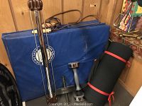 Four blue Everlast folding gym mats stacked against the wall, two weights, bullworker exerciser, black rolled exercise mat with red tie, and tiled padded flooring beneath