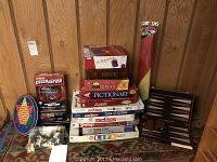 Multiple board games stacked including Monopoly, Pictionary, Stratego, Risk, Catan, Life, Trivial Pursuit, electronic Catch Phrase game with packaging, some loose game pieces and poker chips, Chinese Checkers game board, and backgammon case.