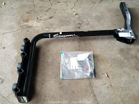 Full view of black metal Swagman bike rack with mounting arm and included hardware in a plastic bag on concrete floor.