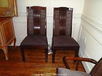 Two dining chairs with dark wood, caned backs featuring central circular emblem, upholstered seats against wall.