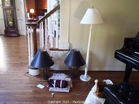 View of two brass lamps with black shades, one white wood floor lamp, and folded fabric items on hardwood floor against stairwell wall.