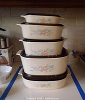 Five nesting ceramic casserole dishes stacked on counter with floral design and brown glass lids.