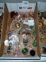 Full view of assorted costume jewelry in cardboard box showing various pieces including necklaces, brooches, rings, earrings.