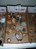 Photo showing a variety of costume jewelry in a box including brooches, earrings, necklaces, and a wristwatch with a blue strap.