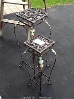 Two metal plant stands with decorative tops and painted leaves on legs, standing side-by-side outdoors on pavement.