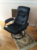 Front angled view of the black leather reclining swivel chair showing tufted backrest, padded armrests, and round metal base on a small rug.