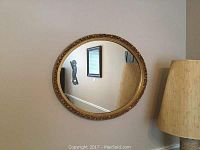 Front view of an oval framed mirror hung on a wall near a floor lamp, showing a gold-tone ornate frame with floral motifs.