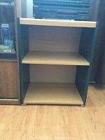 Photo of wooden adjustable shelf unit showing two shelves, black sides, light wood shelves, placed on wooden floor near other furniture.