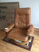 Front view of tan leather recliner chair showing padded armrests, tufted backrest, and round wooden swivel base with metal supports.
