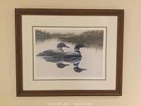 Framed print showing two loons on a lake with their reflections, surrounded by natural foliage, in a brown wood-textured frame.