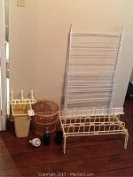 Photo showing assorted bathroom and laundry items including a vertical drying rack, step stool, wicker basket, soap and cup holder, curling iron, and other accessories on wooden floor.