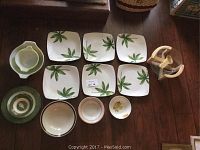 Overview of assorted dishes showing six square dinner plates with green leaf design, assorted small bowls including green round dish, one with floral pattern, and a white salad bowl with serving ladles in a clear and wooden holder.
