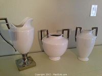 Group photo of three white ceramic vases with silver handles and base on a flat surface against a beige wall.