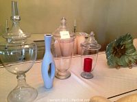 Five piece glass decor lot including three clear apothecary jars with lids, one containing a red candle, one tall blue ceramic vase, and one green decorative art piece shaped like a leaf with a flower accent.