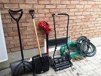 Full lot view showing manual push reel mower, two snow shovels, the Toro cordless trimmer, and two coiled garden hoses against a brick wall.