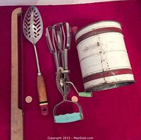All four kitchen tools: wooden bread slicer, slotted spoon, egg beater, and Androck flour sifter on a red background.