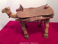 Wooden camel seat with leather cover, showing entire camel figure on red background.