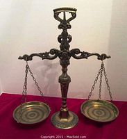 Front view of the brass balance scale on red fabric background showing detail of hanging pans and ornate central arm.