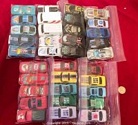 Photo showing multiple small toy cars in clear plastic bags laid out on red fabric background, displaying various colors and designs including blue, white, black, and character-themed cars.