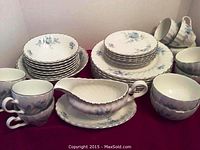 Display of full china set showing stacked dinner plates, bowls, cups, saucers, and gravy boat with drip dish, highlighting the blue floral pattern and silver trim.