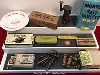 Photo showing full assortment of vintage miscellaneous items arranged on trays including two metal boxes, sets of pen nibs, brass thumbtacks, old keys on a key ring, miniature stove, salt tablets, and ceramic hotel dish.