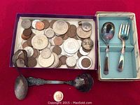 Top view of a box containing assorted silver and silver-plated coins alongside a small box containing a child's silver-plated fork and spoon on a red background.