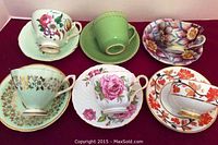 Front view of all six tea cup and saucer sets arranged in two rows against a pink background showing varied floral and solid designs