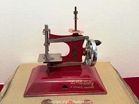 Front view showing the full red toy sewing machine with decorative gold design and hand wheel with black handle.
