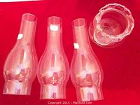 Photo of three clear glass oil lamp chimneys and one clear glass vase with scalloped rim placed on a red fabric background.
